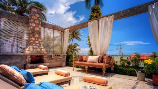 A sunlit outdoor seating area with a stone fireplace, a cushioned sofa, and a bench. - Villas Enrique (Puerto Vallarta)