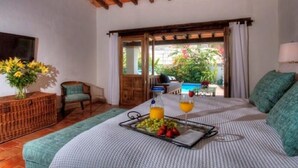 A cozy room with a wooden ceiling, a bed with a striped bedspread, a small table with fruit and drinks, a wicker side table, and a view of a garden through a glass door. - Villas Enrique (Puerto Vallarta)