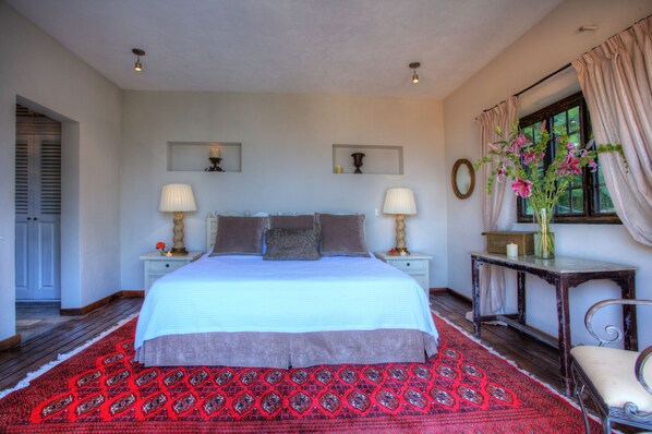 A bedroom with a large bed, two bedside tables with lamps, a nightstand, a chair, a window with curtains, and a red patterned rug.