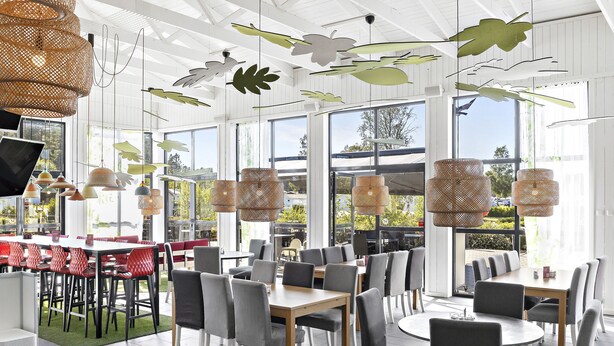 A modern dining area with multiple tables and chairs, large windows, and hanging wicker lamps. - First Camp Karlstad (Karlstad)