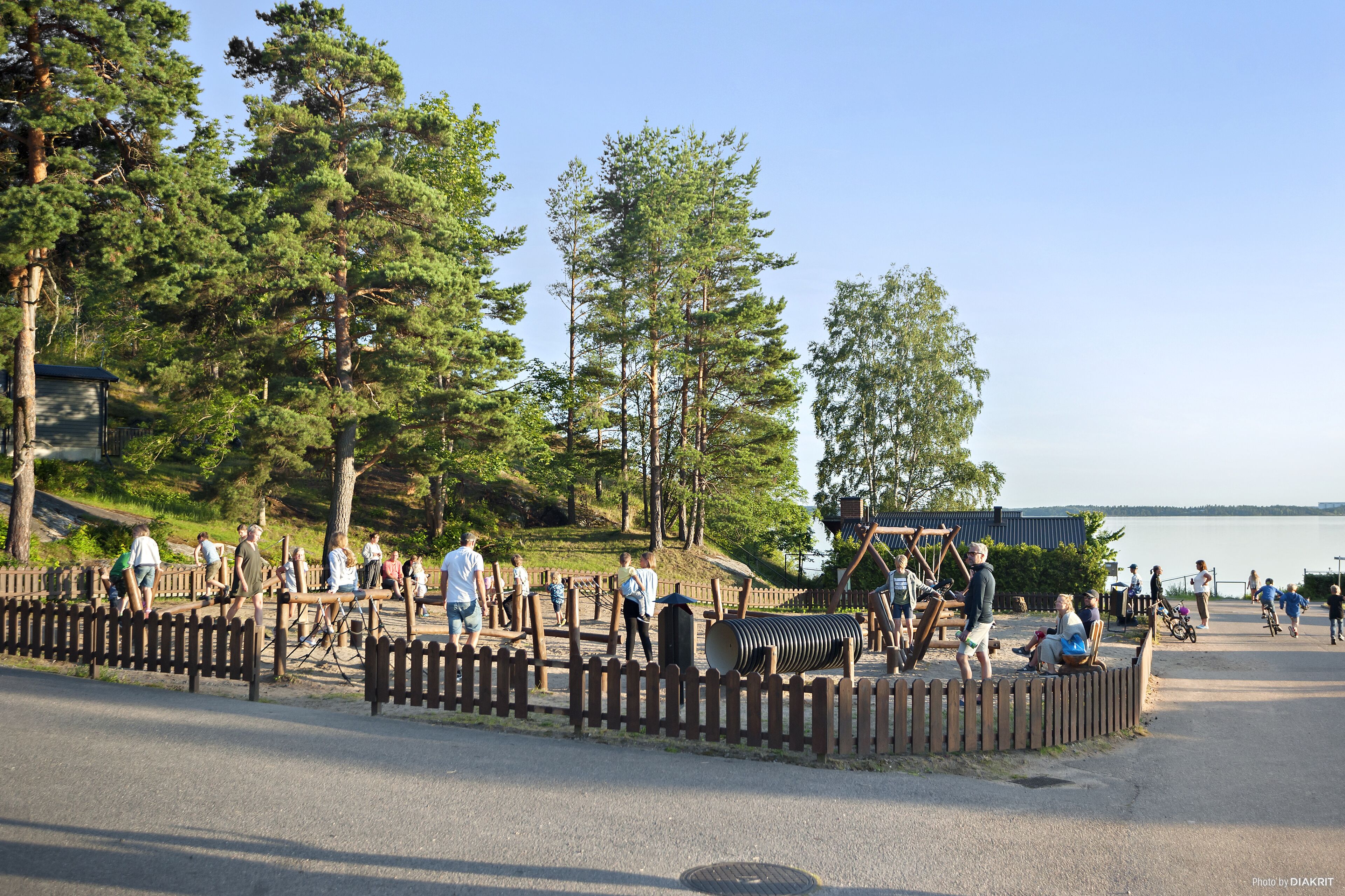 Un parque con valla de madera, gente caminando y un lago al fondo.