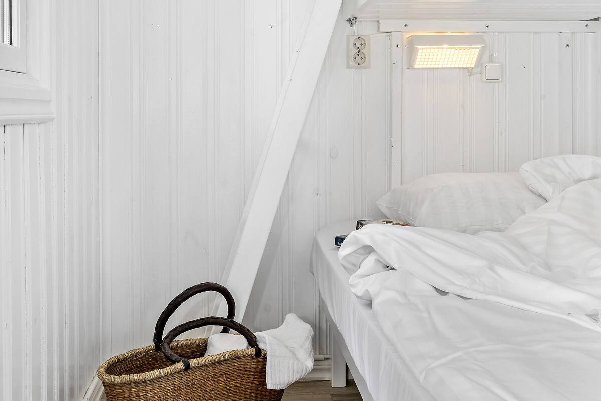 Een kleine, witte kamer met een enkel bed, schuine plafonds, een houten vloer en een rieten mand bij de ingang.
