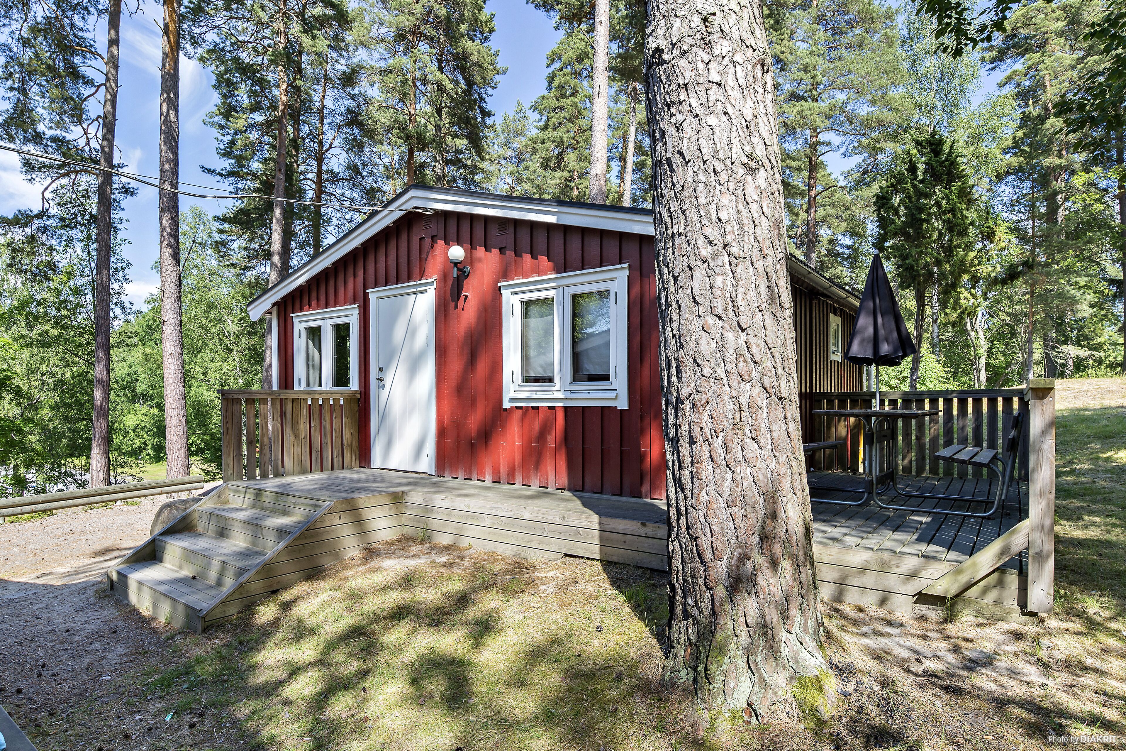 Une petite cabane rouge avec une terrasse en bois et un espace salon abrité, entourée de grands arbres.