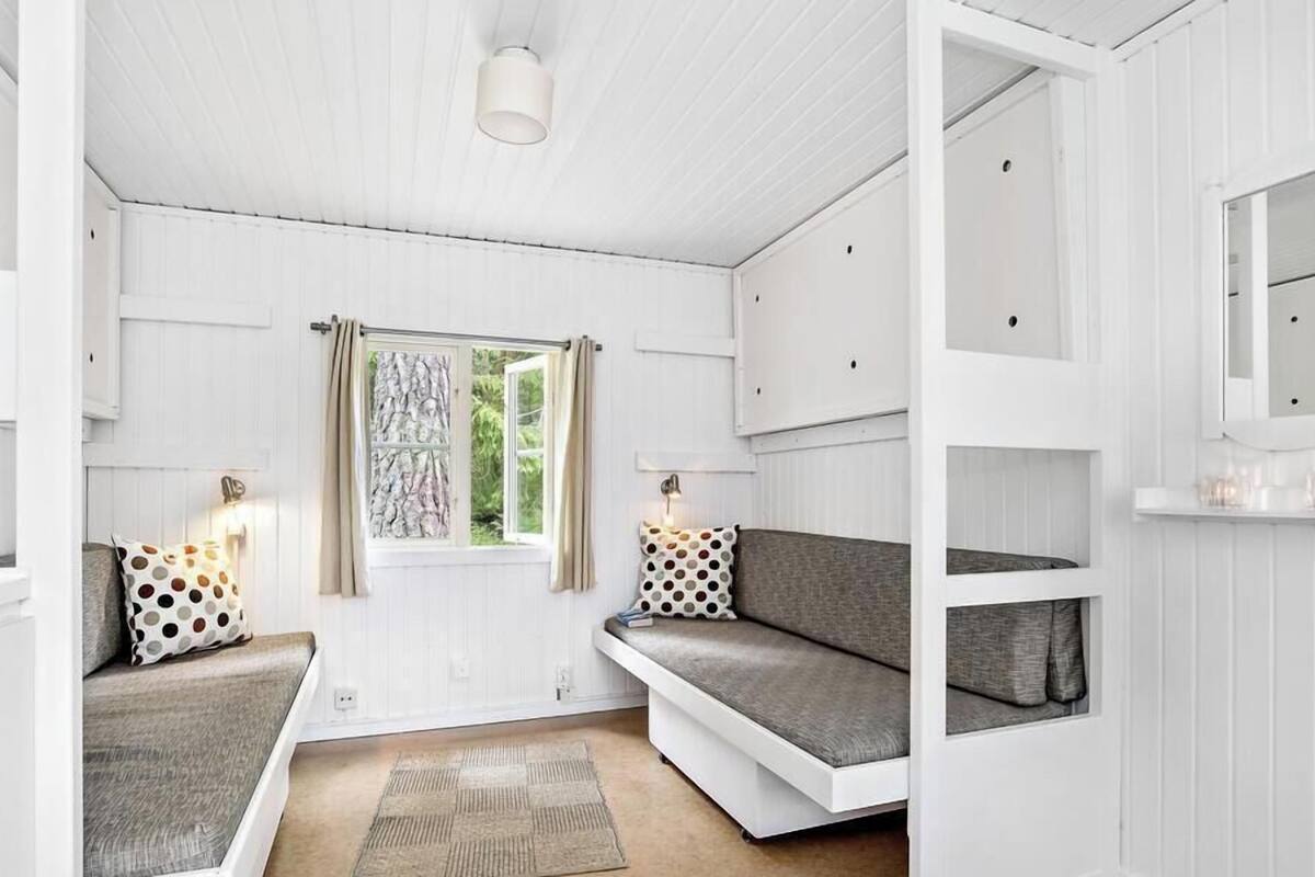 Een kamer met een witte inrichting, een raam met gordijnen, een bank met een stippen kussen, een tafeltje met een lamp en een spiegel aan de muur.