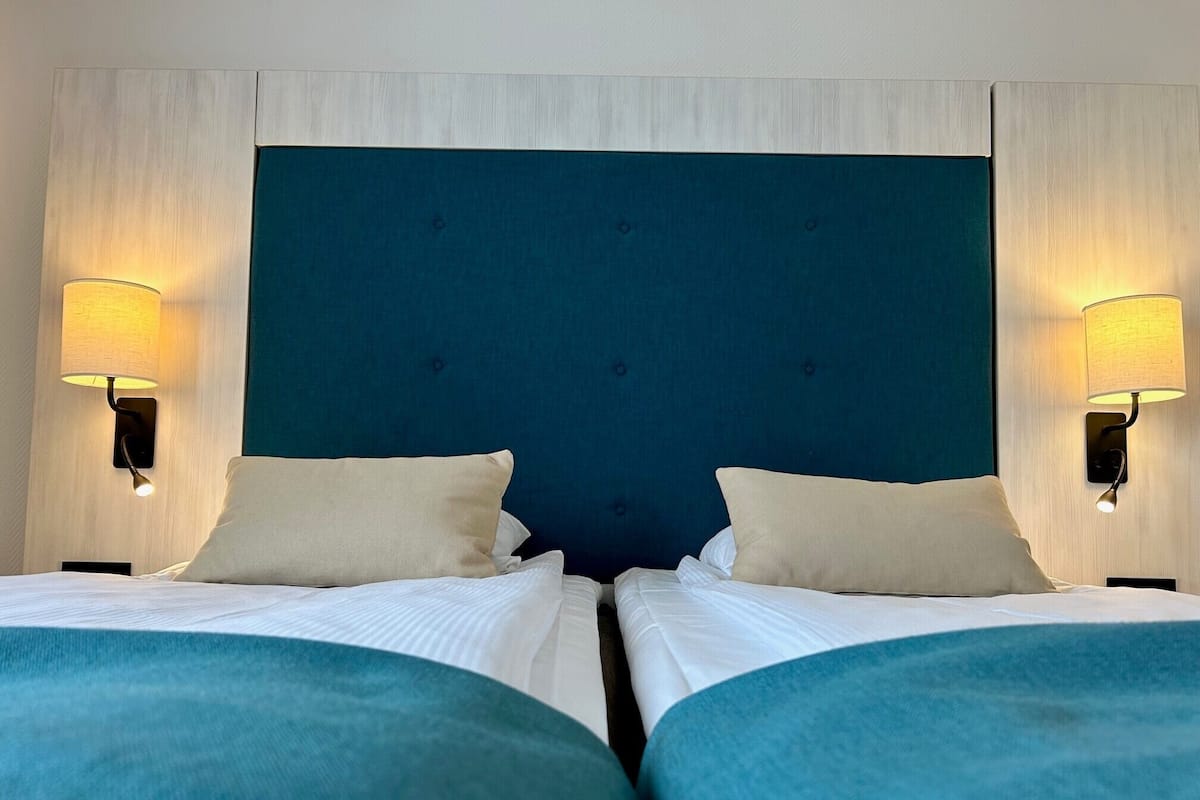 A hotel room with two beds, each with a blue headboard and white bedding. There are two wall-mounted lamps on either side of the beds.
