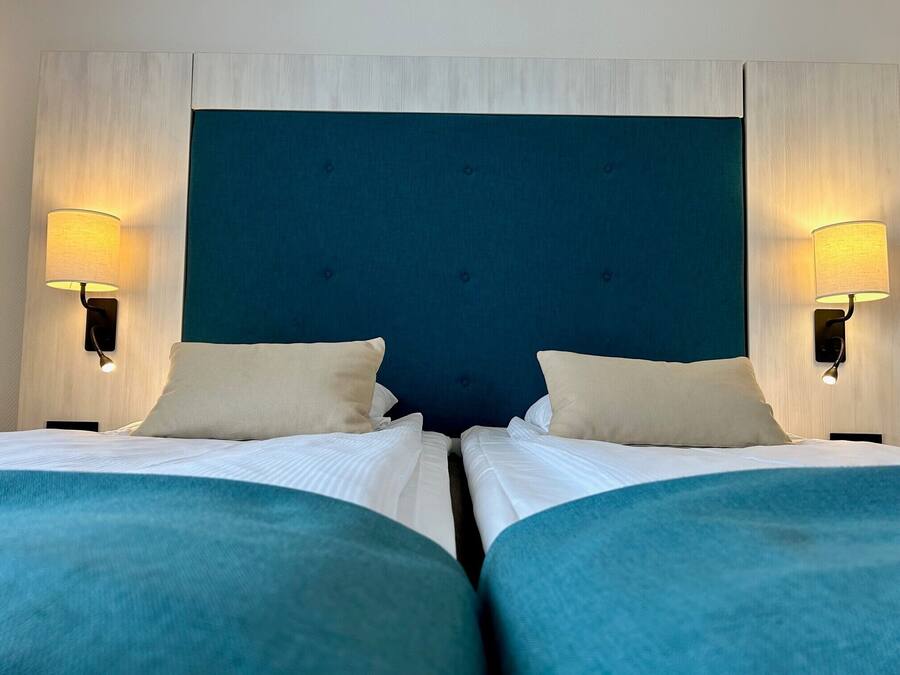 Habitación de hotel con dos camas, cada una con cabecero azul y ropa de cama blanca. Hay dos lámparas fijadas a la pared a cada lado de las camas.