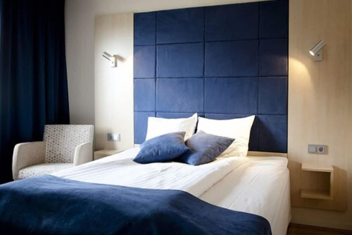 A modern hotel room with a large bed, a headboard with a padded design, a bedside table, and a chair.