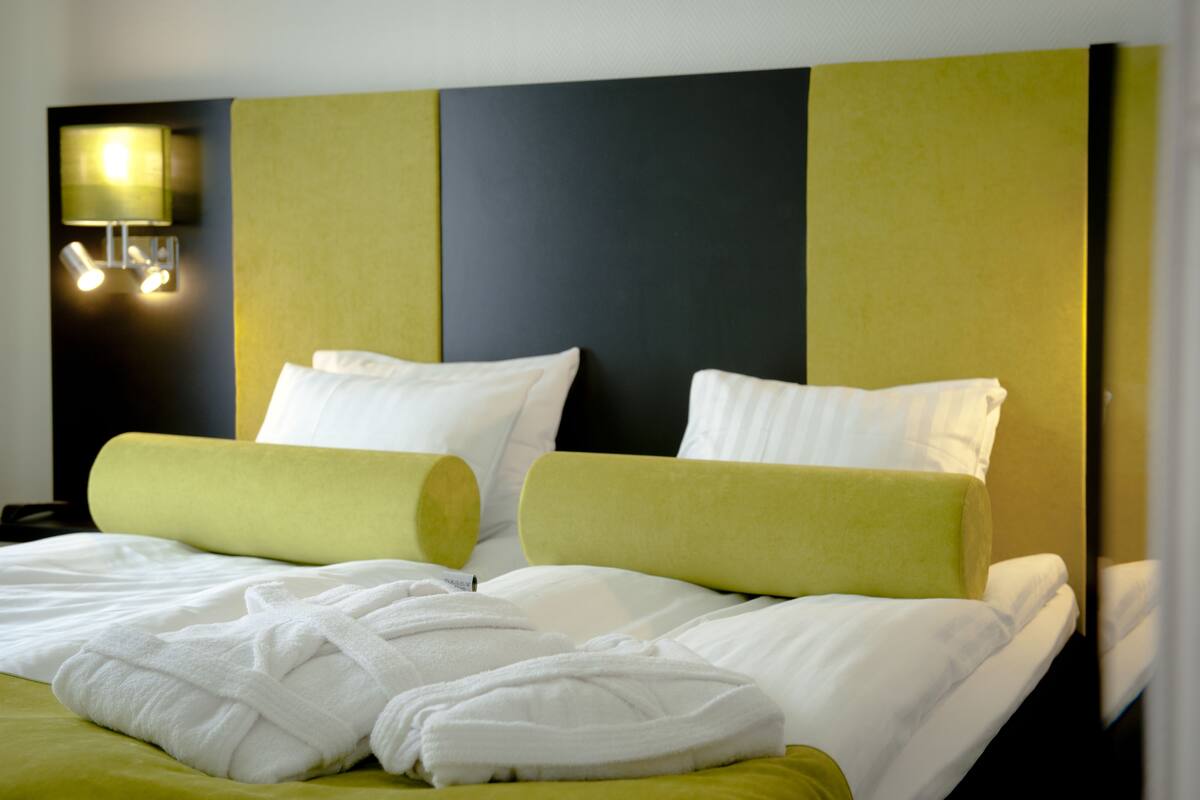 A hotel room with a large bed, two pillows, a nightstand, and a wall-mounted lamp.
