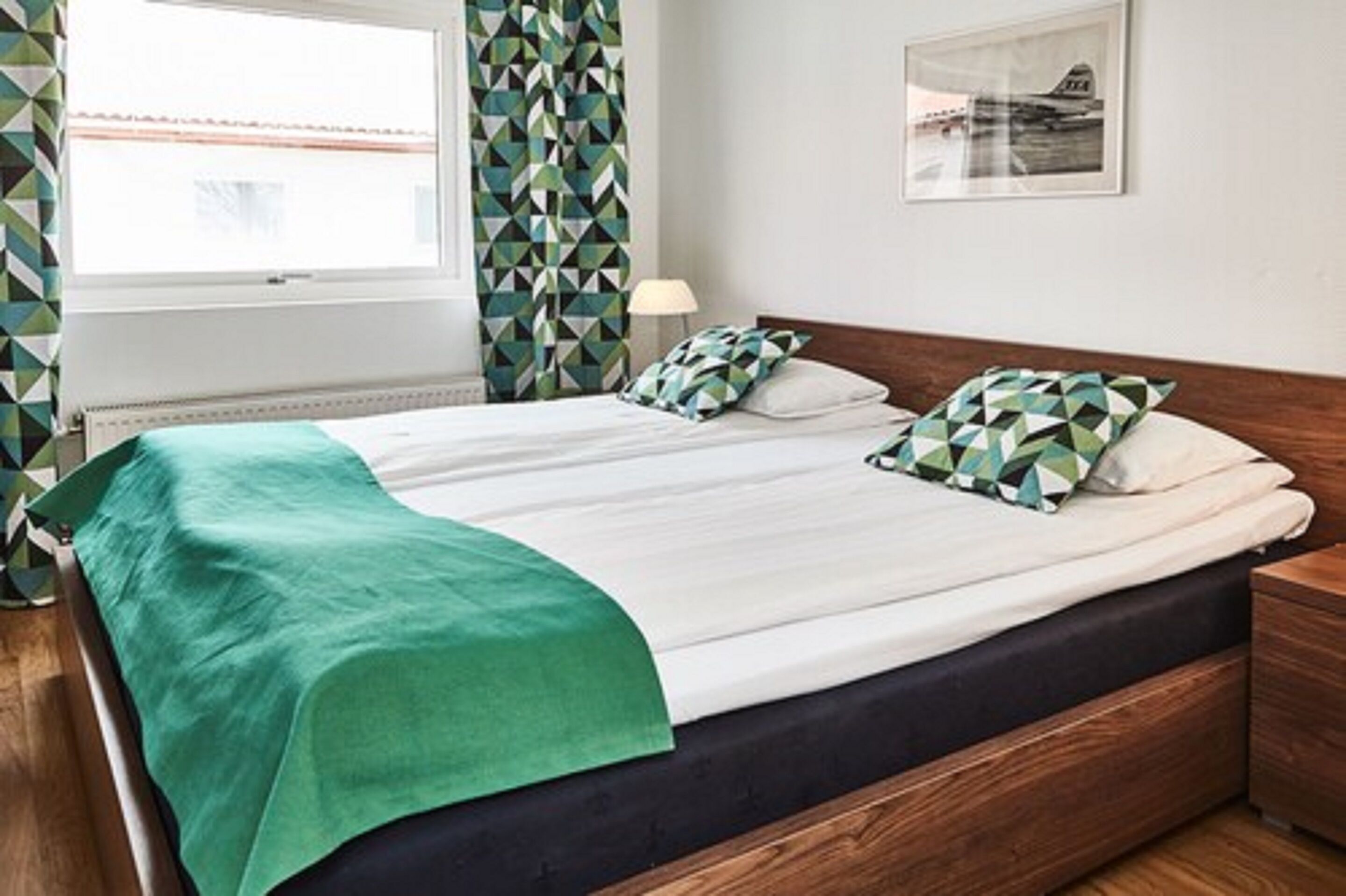 A neatly made bed with a green throw, a wooden headboard, and a nightstand with a lamp.