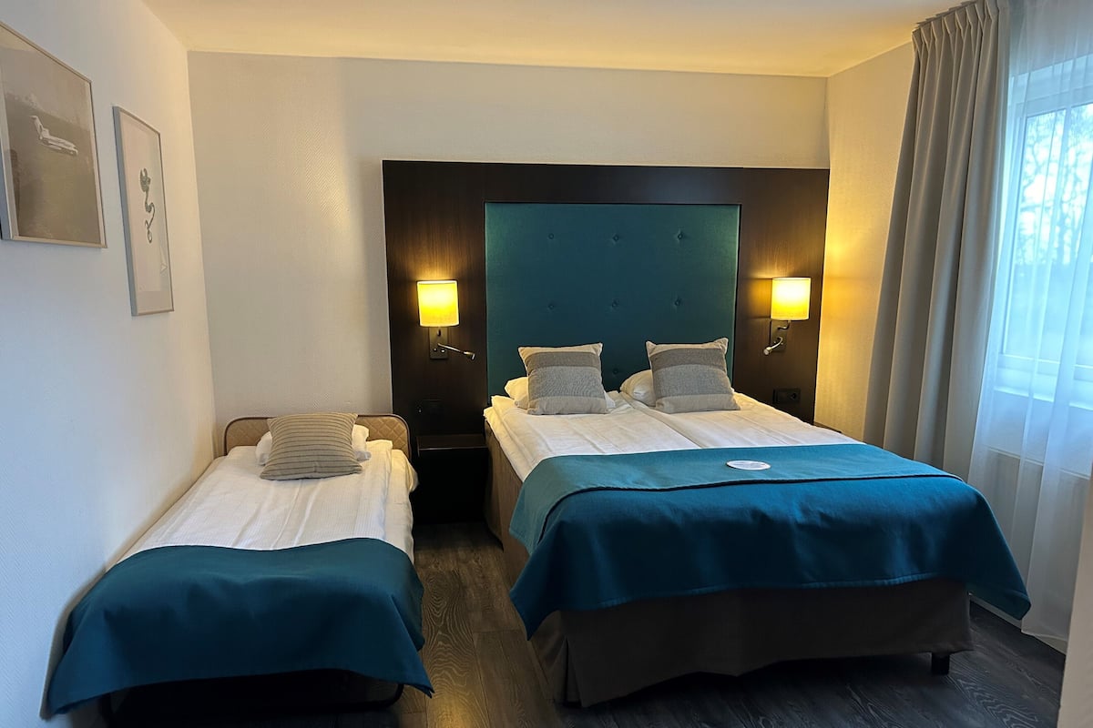 A hotel room with two single beds, a dark headboard, bedside lamps, and a window with curtains.