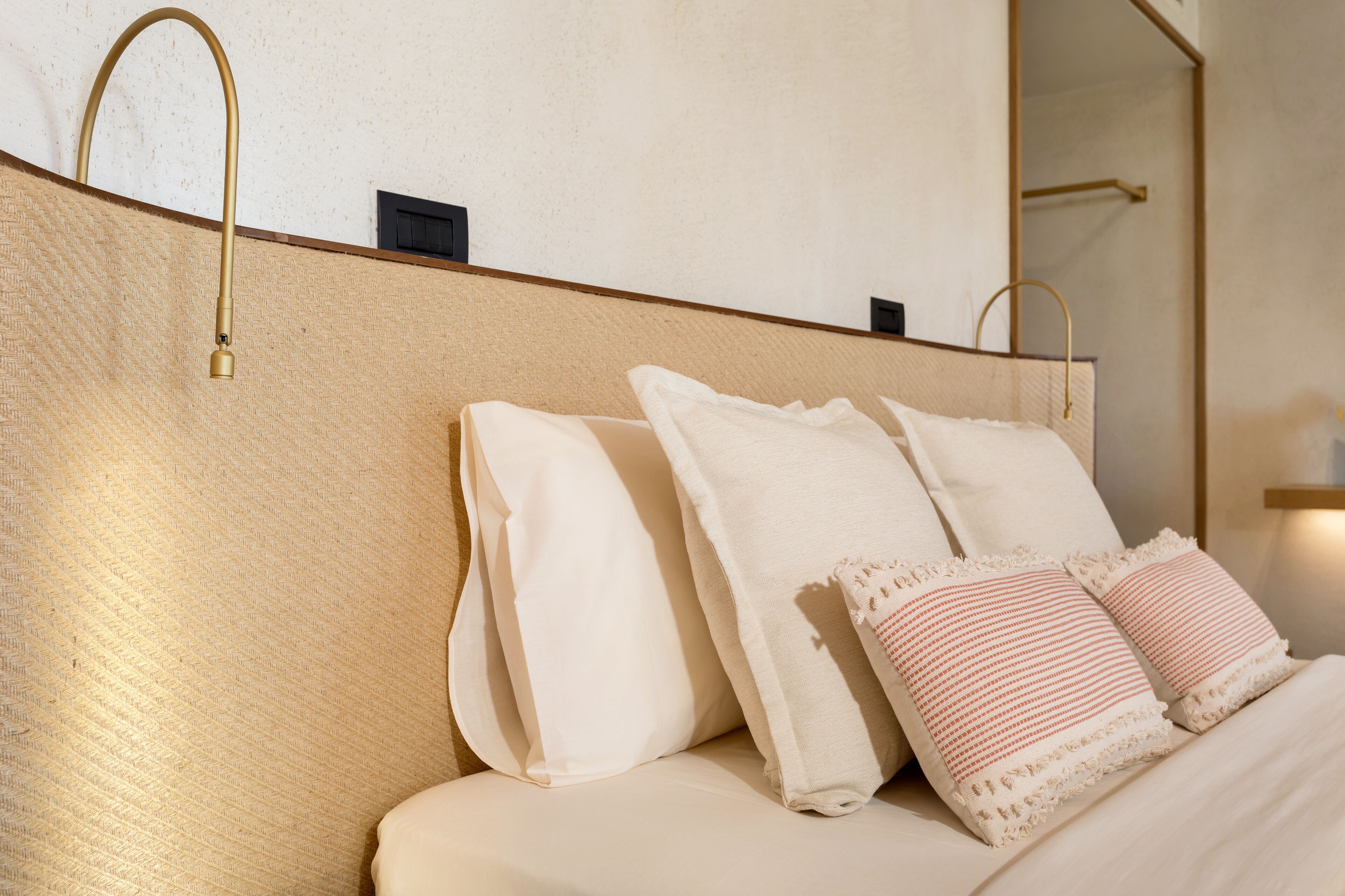 Una cama bien hecha con almohadas blancas y rosas, un cabecero con diseño texturizado y un grifo dorado en la pared.