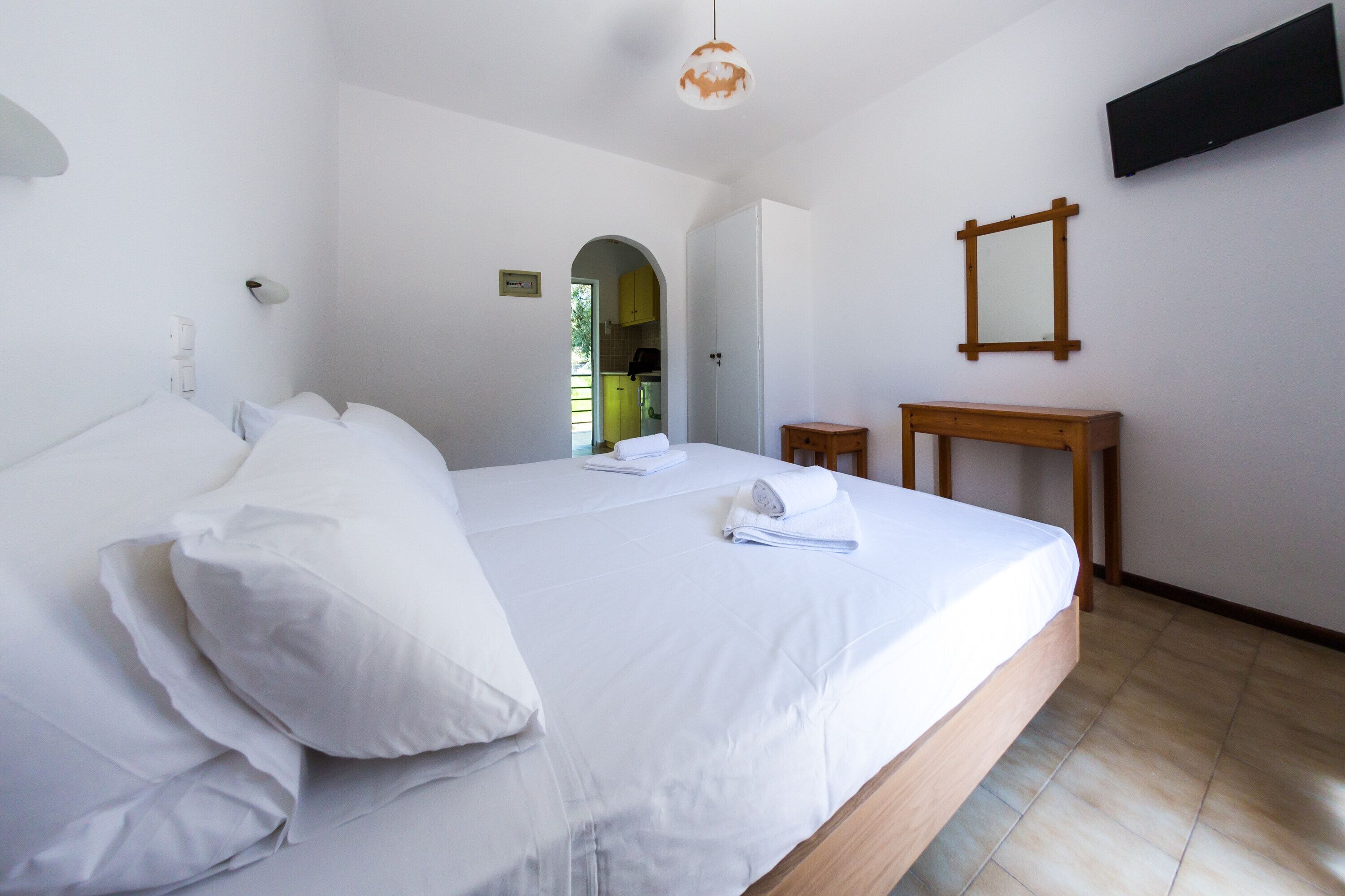 A neatly made bed with white linens, a wooden bedside table, a small mirror, and a television mounted on the wall.