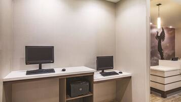 A modern office space with two desks, each equipped with a computer monitor, keyboard, and mouse. There is a printer on the left desk. The room has a neutral color scheme with a light-colored wall and a wooden floor.