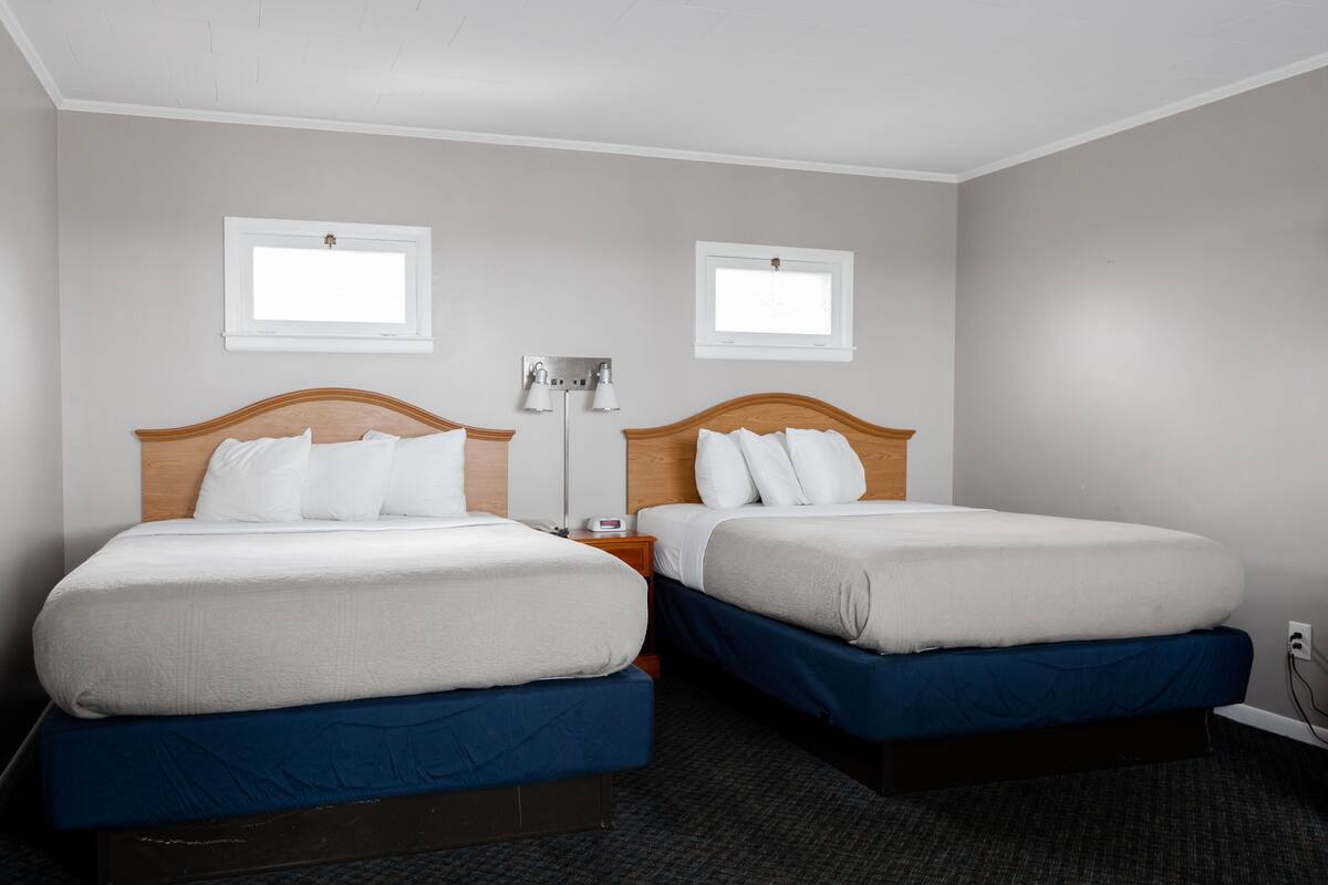 A hotel room with two beds, a nightstand, a lamp, and two windows.
