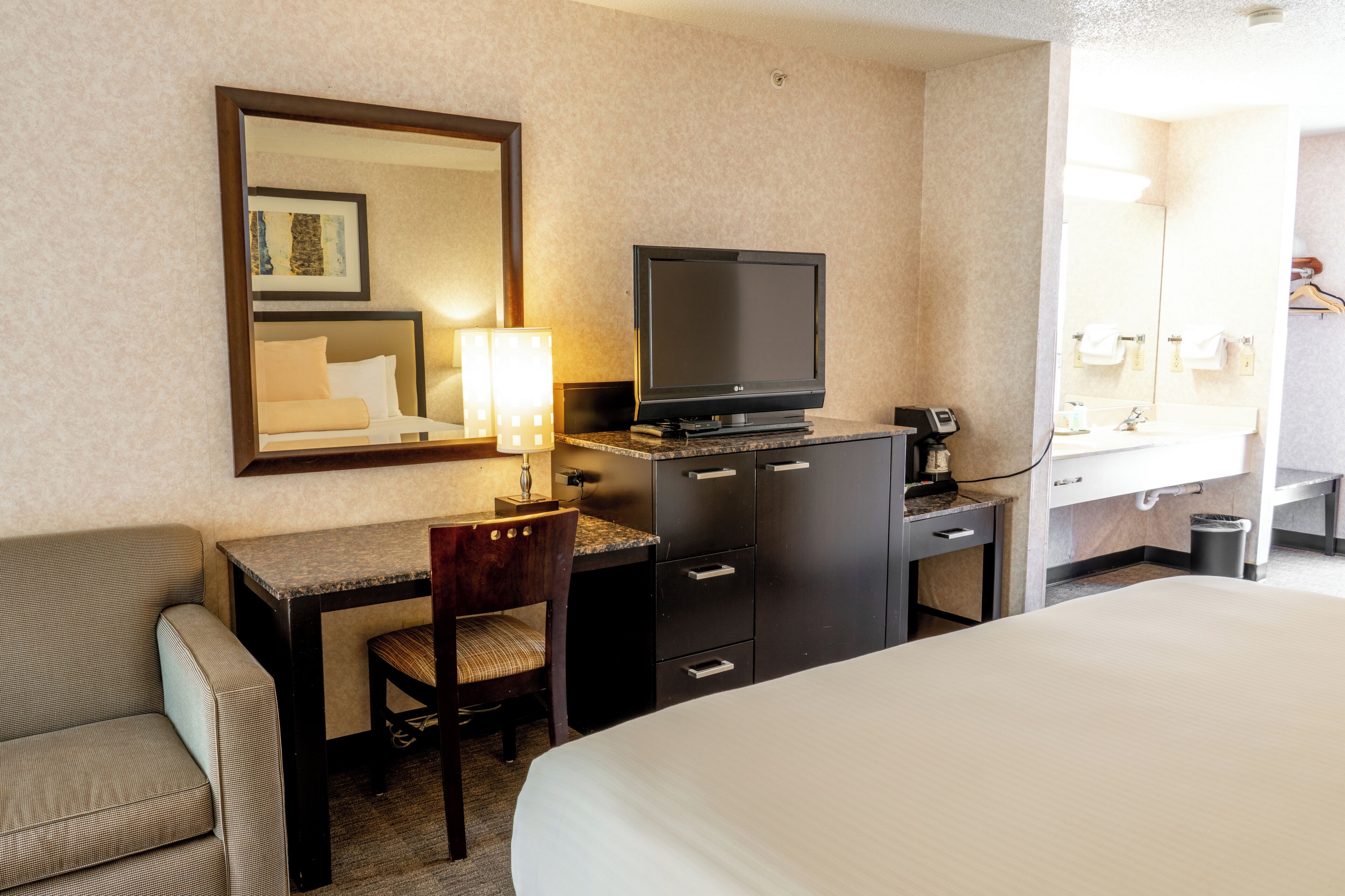 A hotel room with a bed, a desk with a chair, a television, a mirror, and a bathroom with a sink and towel rack.