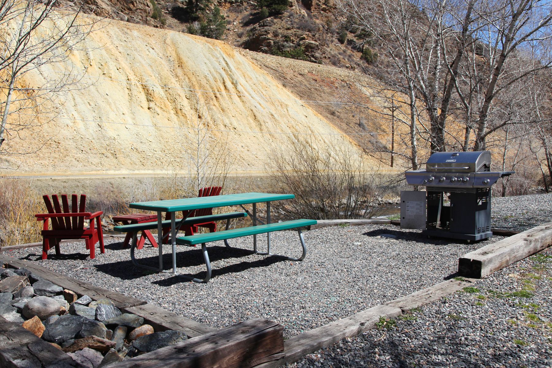 Un espace pique-nique avec des tables et des chaises de pique-nique rouges et vertes, un barbecue et une colline rocheuse en arrière-plan.