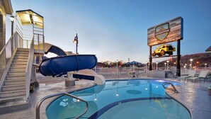 Un espace piscine avec un toboggan, un panneau indiquant « Expedition Lodge », et un espace extérieur aménagé avec des sièges, le tout délimité par une clôture.