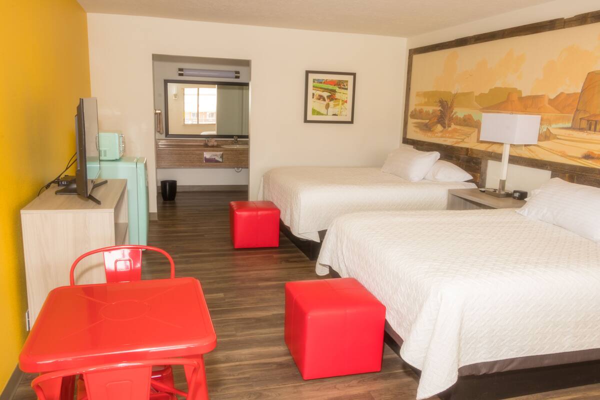 A hotel room with two beds, a desk with a computer, a red chair, and a red ottoman.