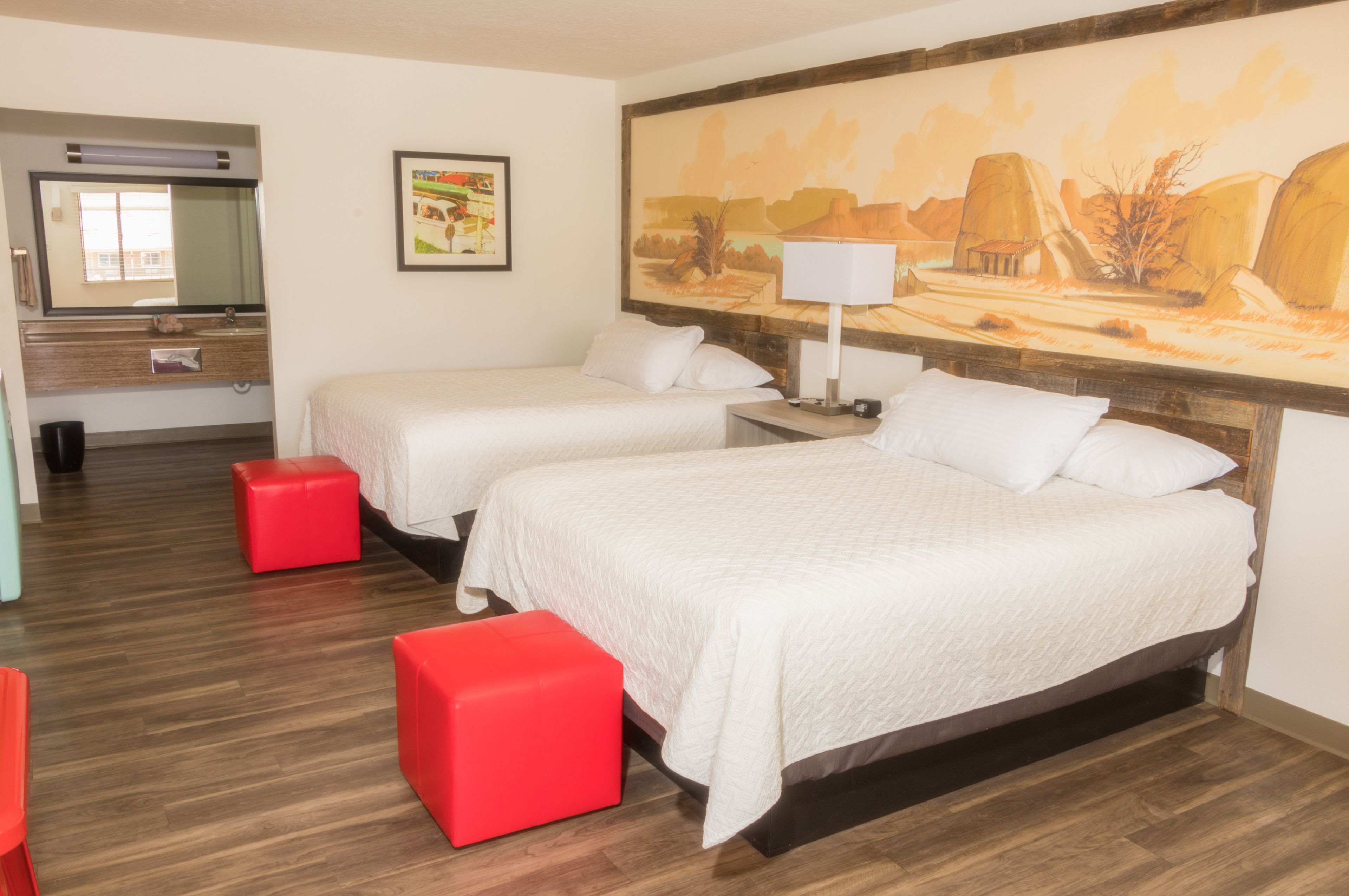 Une chambre d’hôtel avec deux lits, un pouf rouge, un tableau au mur et un plancher en bois.