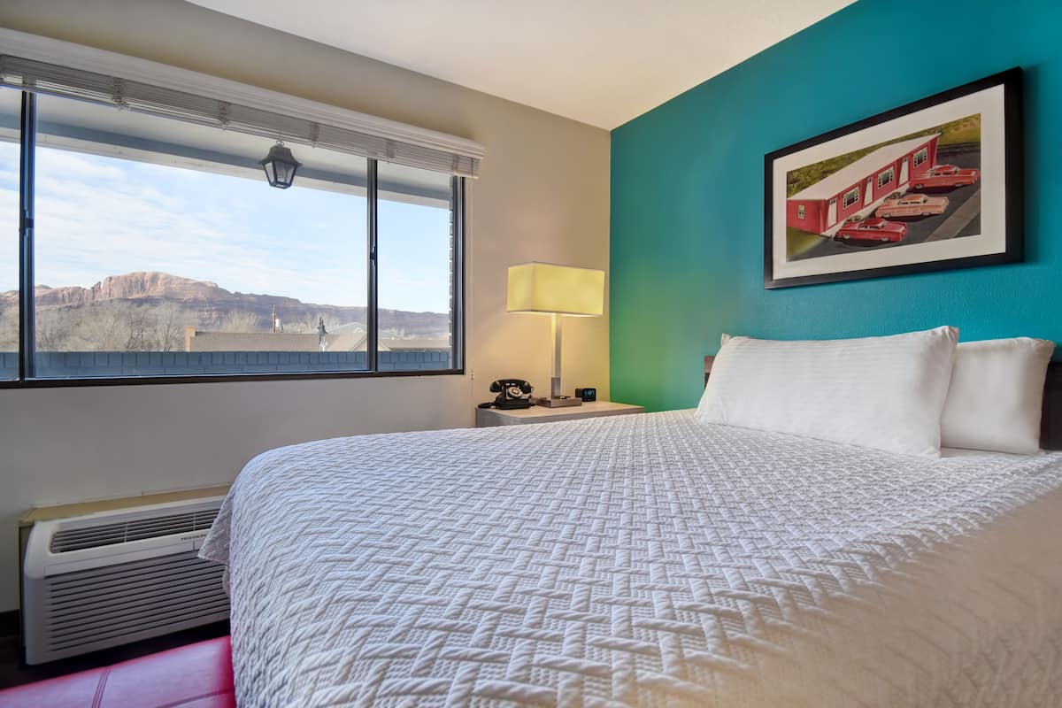 A hotel room with a large bed, a window with a view of mountains, a lamp, and a framed picture on the wall.