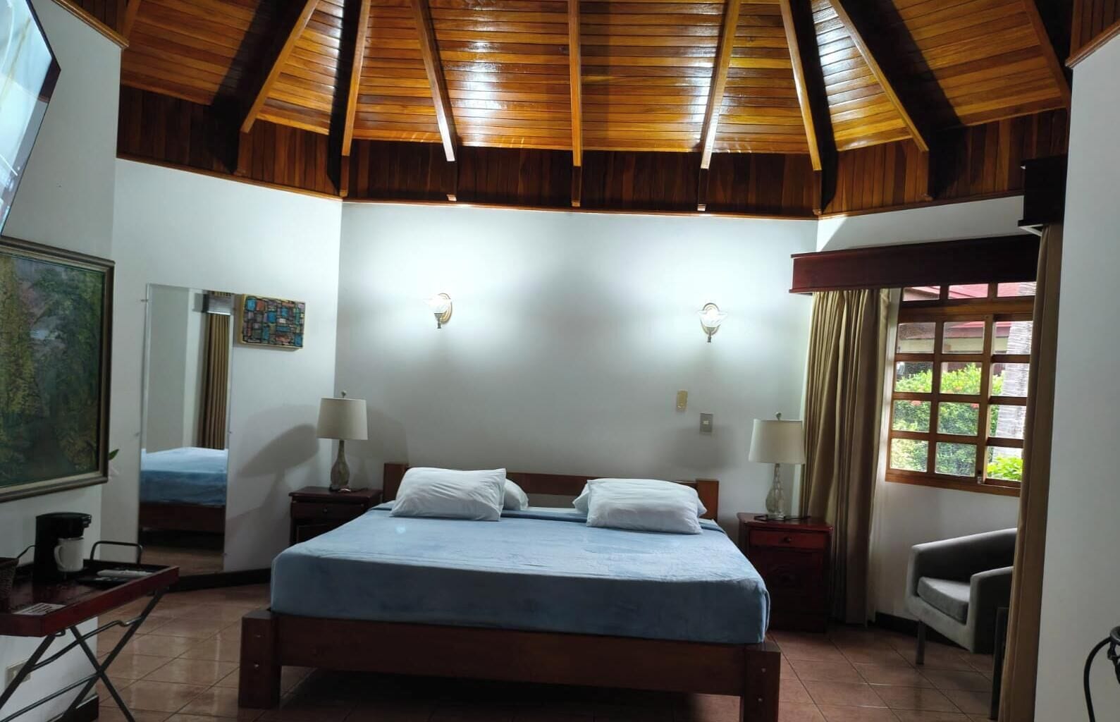 A bedroom with a wooden ceiling, a bed, bedside tables, a chair, a window with curtains, and a painting on the wall.