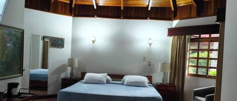 A bedroom with a wooden ceiling, a bed, bedside tables, a chair, a window with curtains, and a painting on the wall.