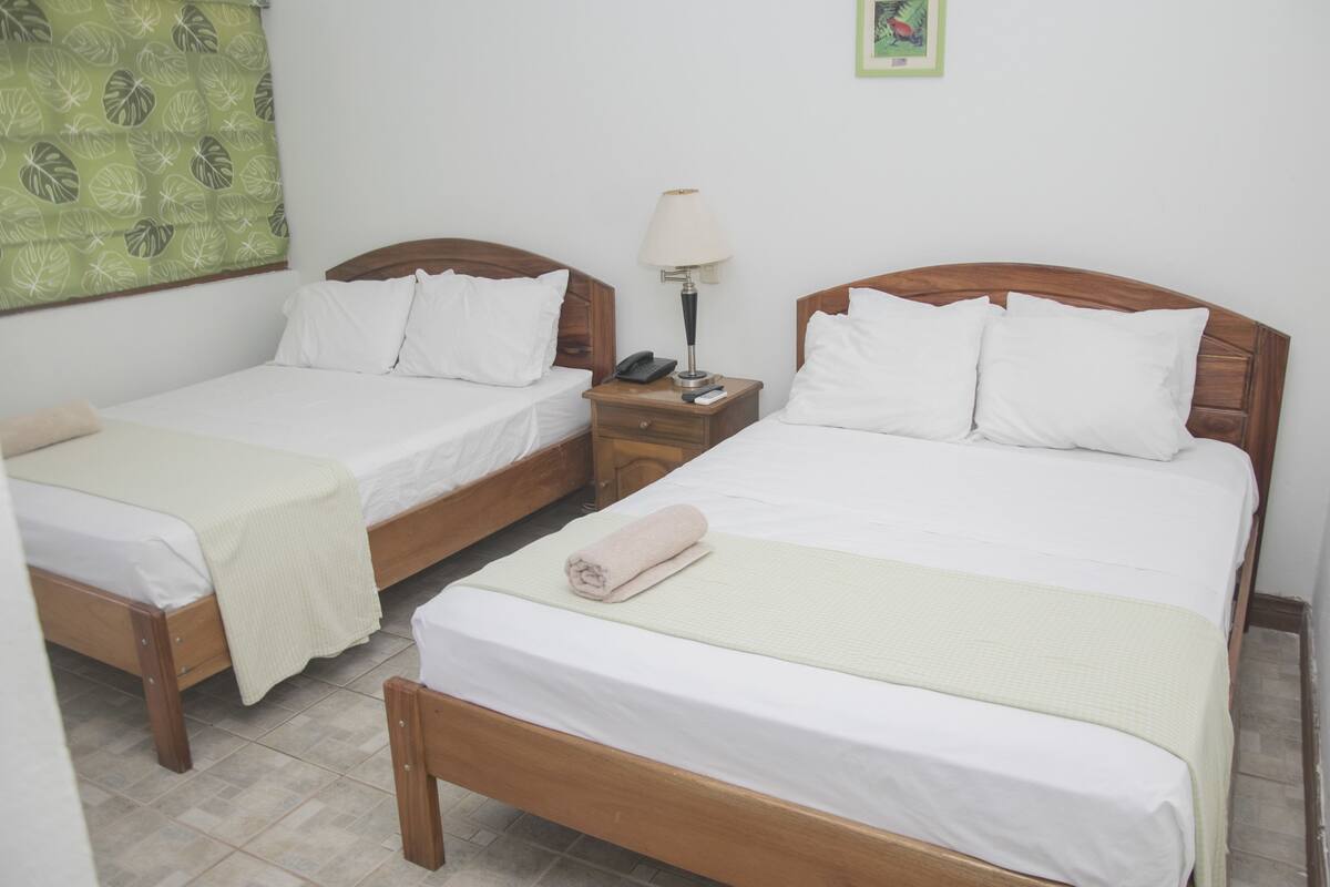 A hotel room with two single beds, a nightstand with a lamp, and a small table.