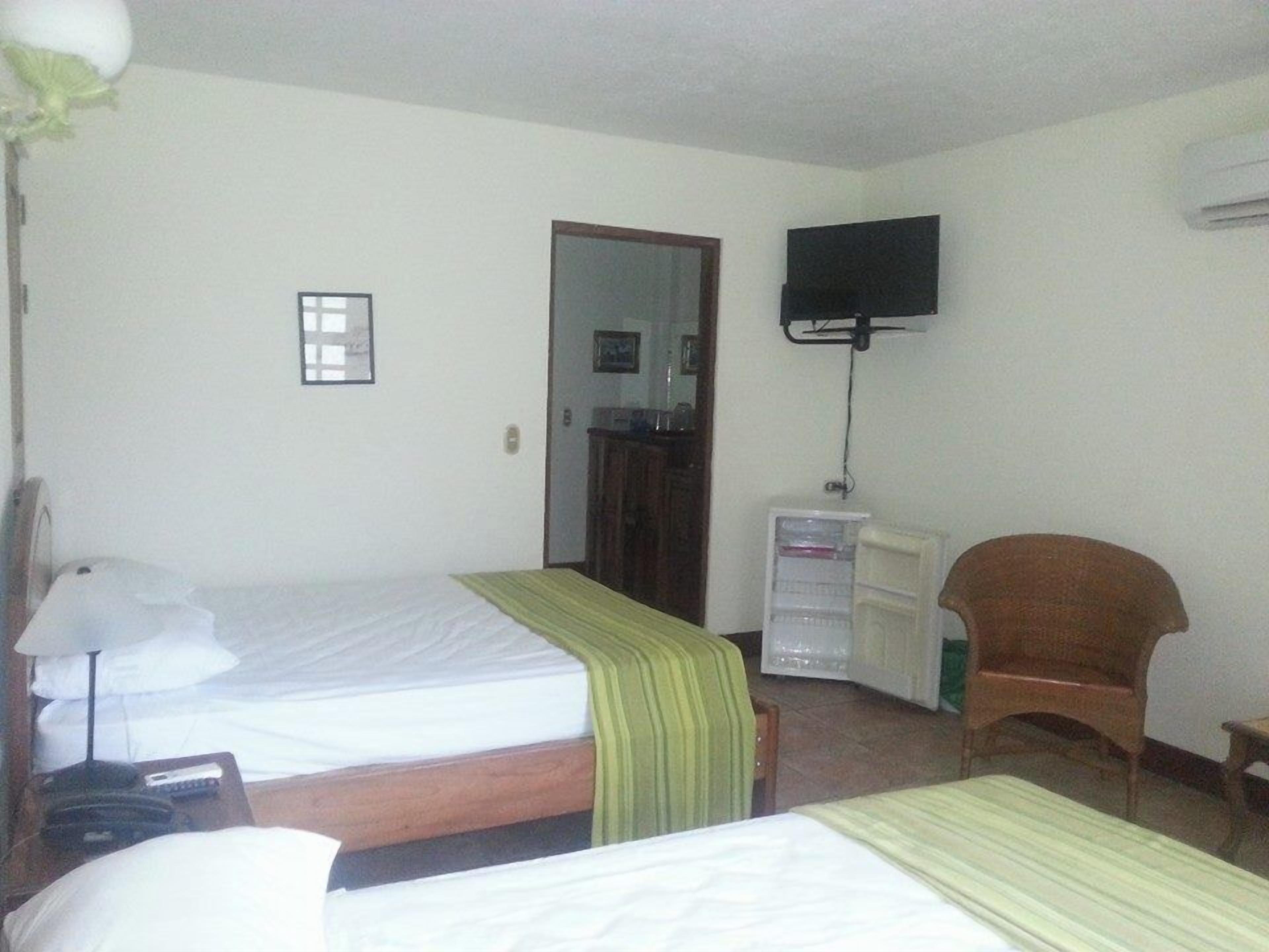 Quarto de hotel com duas camas, uma televisão fixada na parede, uma mesa de trabalho com uma cadeira e um guarda-roupa.