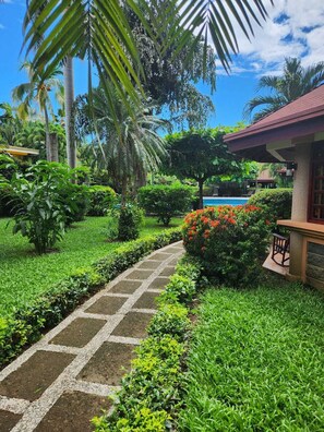 A garden pathway leading to a building with a pool, surrounded by lush greenery and palm trees. - Hotel Villa Acacia (Sardinal)