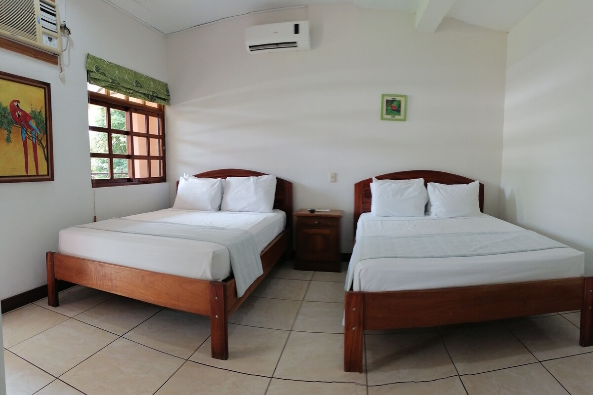 A hotel room with two wooden beds, a small bedside table, a window with a green curtain, and an air conditioning unit on the wall.