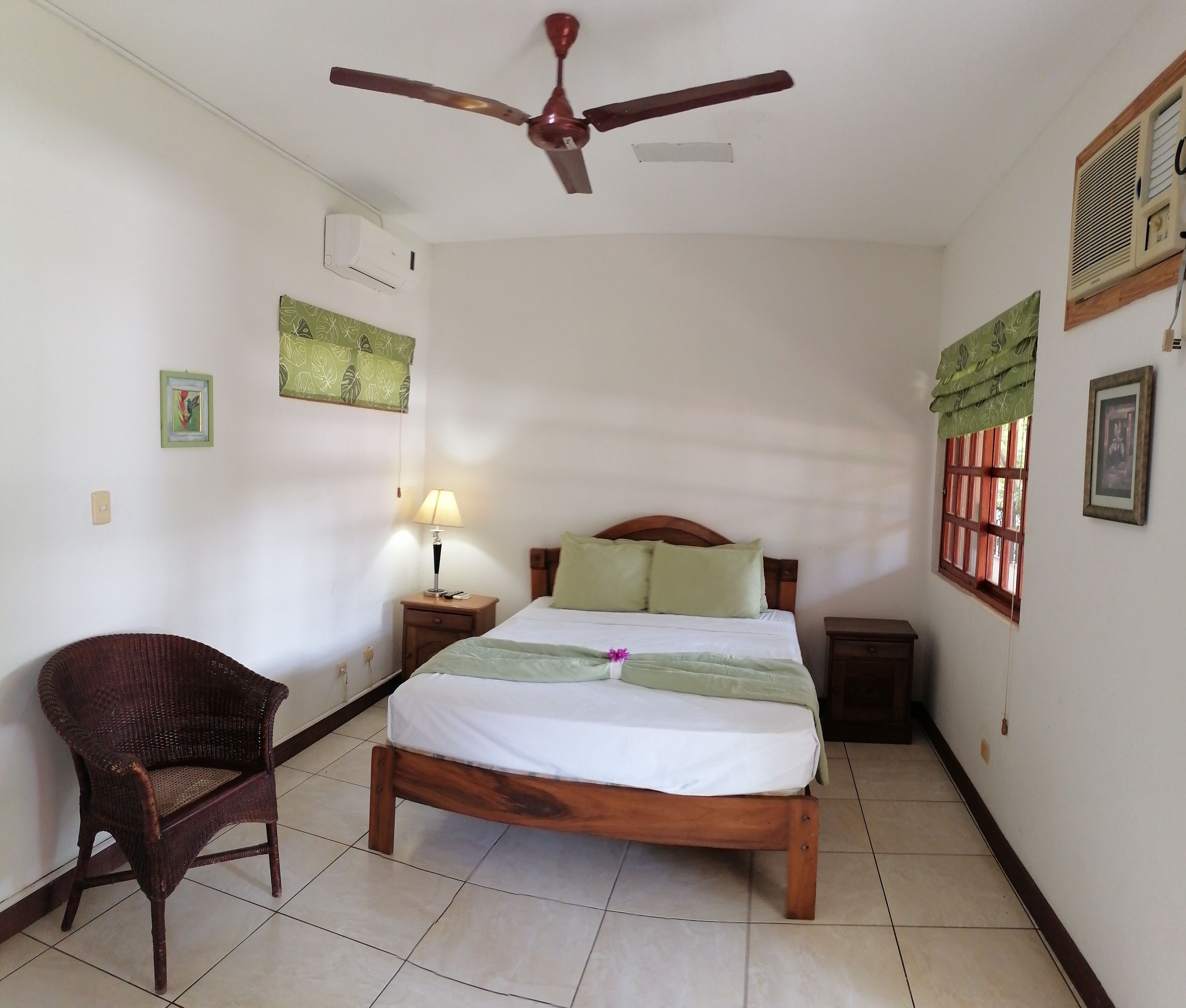 A bedroom with a bed, a chair, a bedside table, a lamp, a ceiling fan, and a window with shutters.