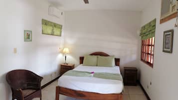 A bedroom with a bed, a chair, a bedside table, a lamp, a ceiling fan, and a window with shutters.