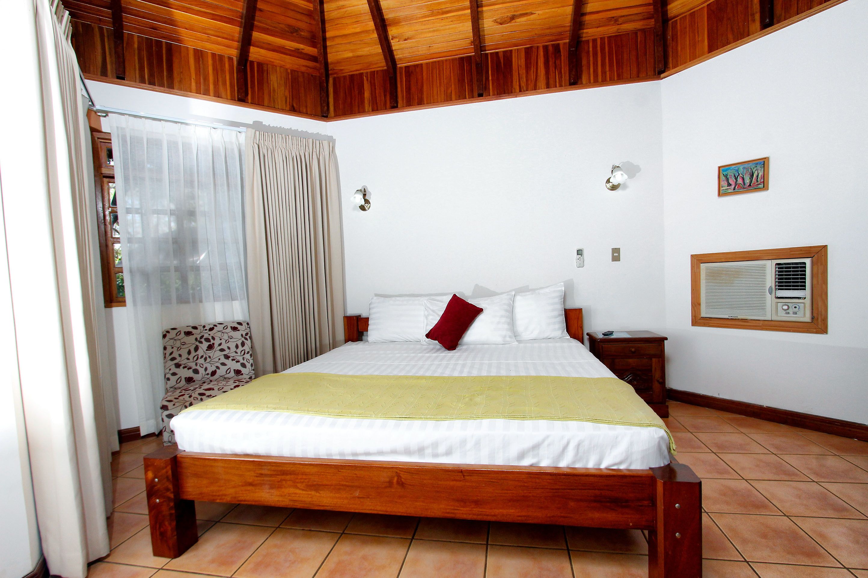 A bedroom with a wooden bed, white bedding, a yellow throw, a red pillow, a nightstand, a chair, a window with blinds, and a wall-mounted air conditioning unit.