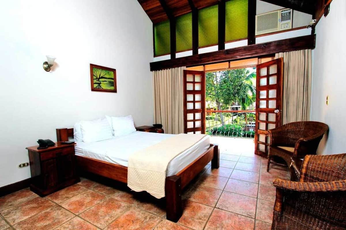 A bedroom with a wooden bed, a nightstand, a wicker chair, and a balcony with a view of greenery.