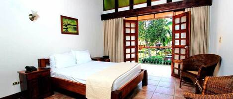 A bedroom with a wooden bed, a nightstand, a wicker chair, and a balcony with a view of greenery.
