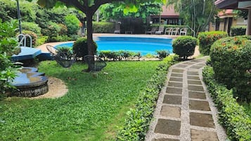 Una piscina con un camino de piedra que la conduce, rodeada de abundante vegetación y un árbol que proporciona sombra.