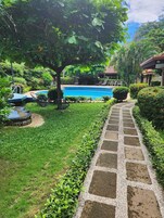 Una piscina con un camino de piedra que la conduce, rodeada de abundante vegetación y un árbol que proporciona sombra.