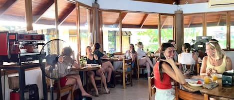 A communal dining area with wooden tables and chairs, a fan, and a coffee machine.