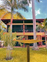 Un edificio de dos pisos con balcón, rodeado de palmeras y una hamaca.