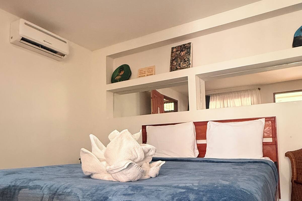 A bedroom with a bed, a shelf with decorative items, a window with curtains, and an air conditioning unit.