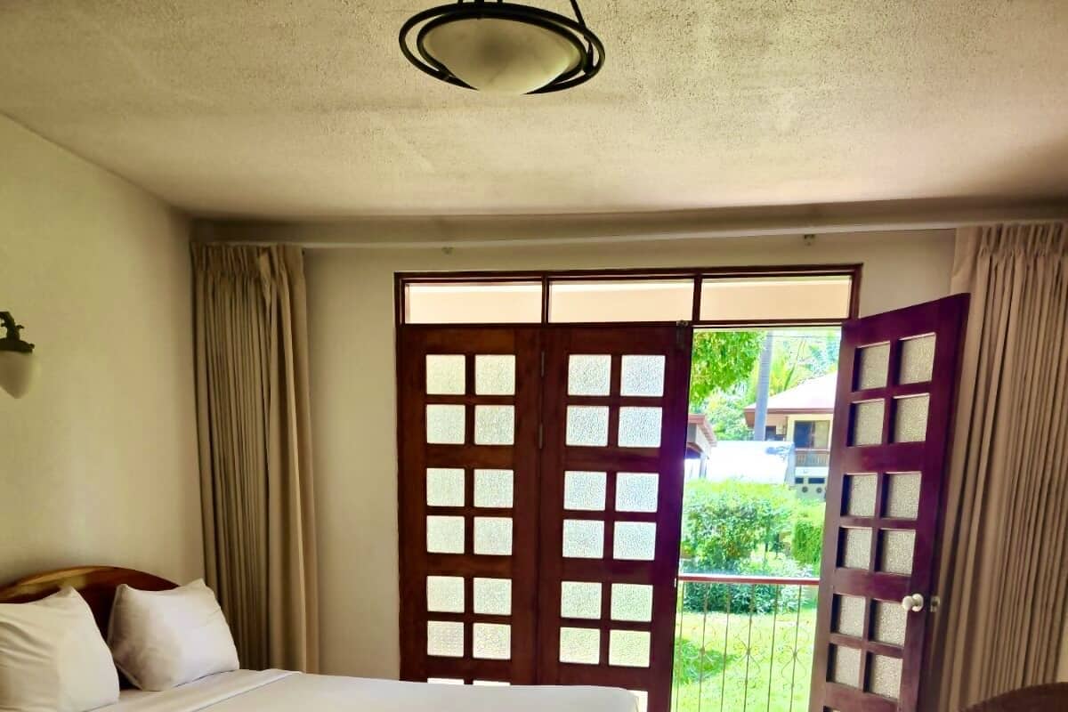 Ein Hotelzimmer mit zwei Betten, einem Sessel, einem Balkon mit Blick auf Grünflächen und einer Tür, die nach draußen führt.