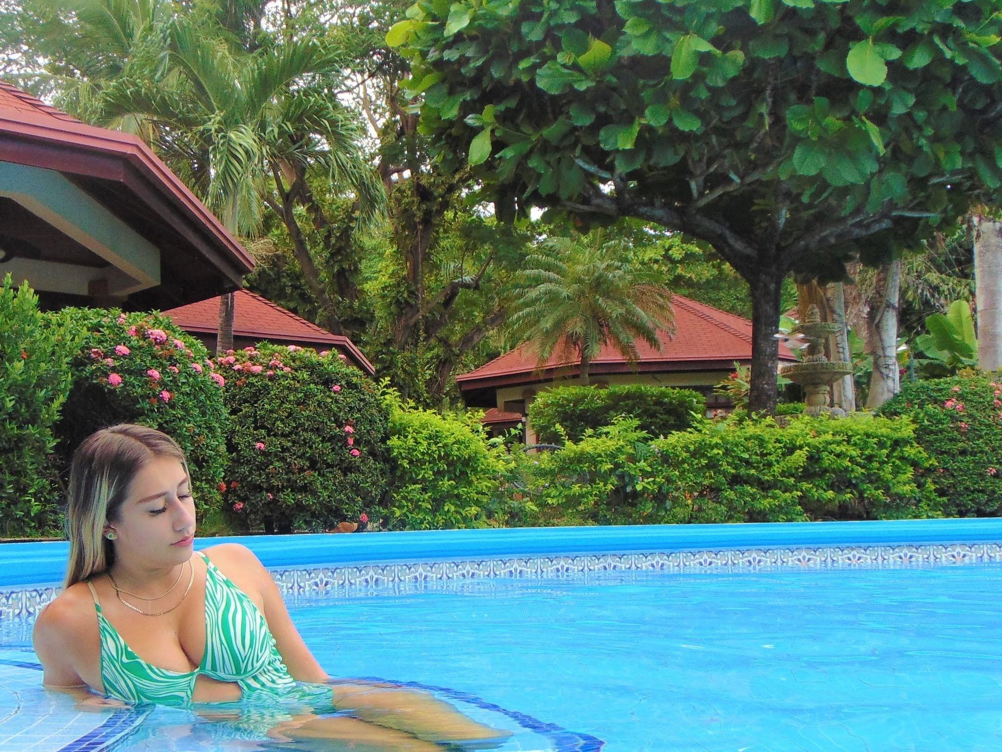 A swimming pool with a tiled edge, surrounded by lush greenery and a building with a red roof.