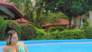 Una piscina con borde de azulejos, rodeada de exuberante vegetación y un edificio de techo rojo.