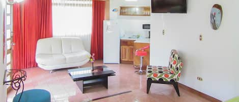 Una sala de estar con un sofá blanco, un sillón estampado, una mesa de centro y un televisor montado en la pared.