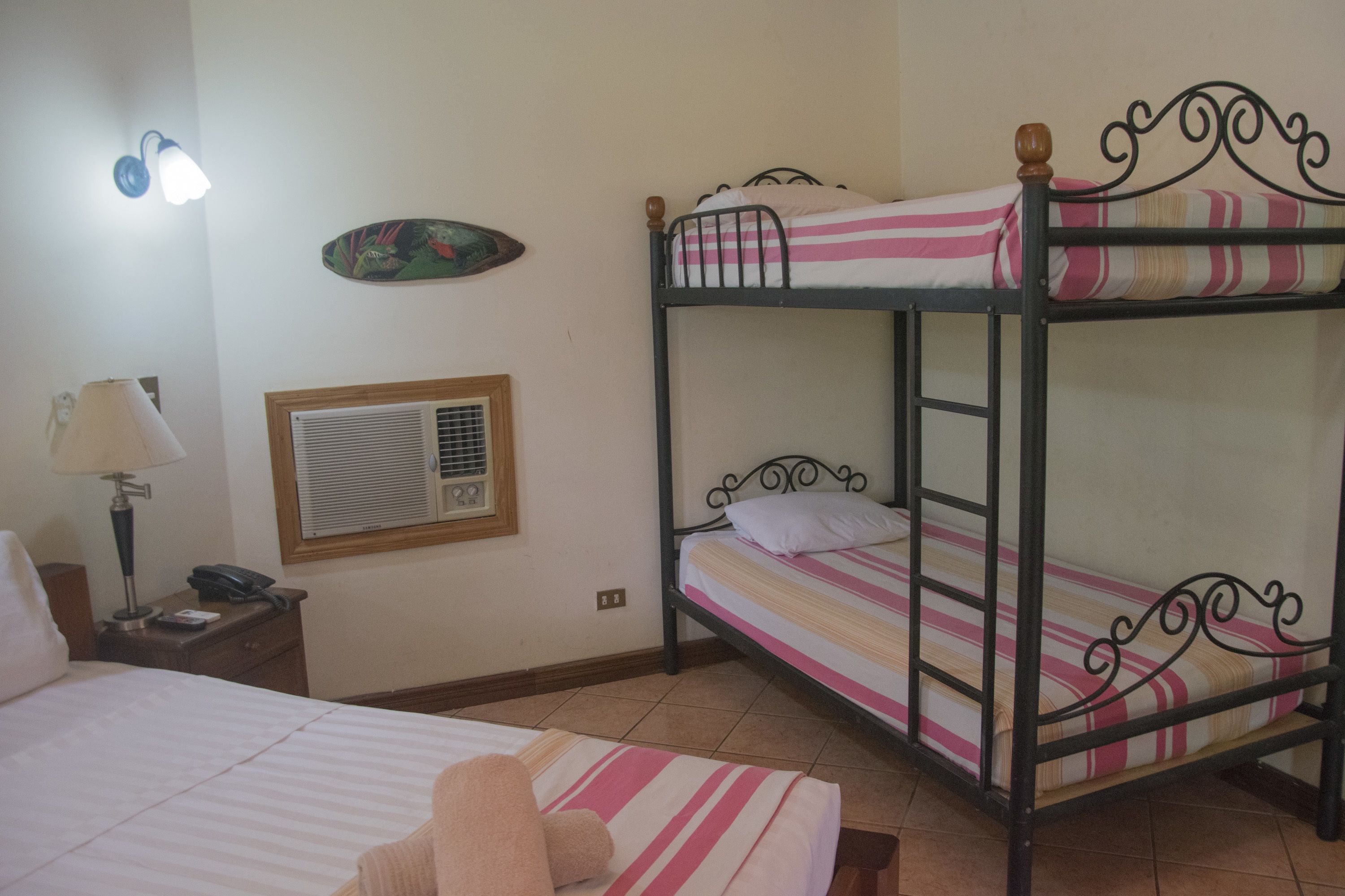 Um quarto com uma cama beliche, uma cama de solteiro, uma mesa de cabeceira com abajur, um relógio de parede e um prato decorativo.