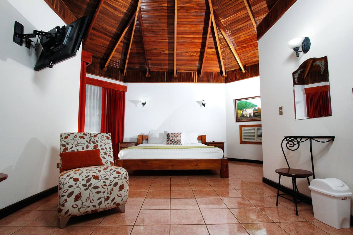 A bedroom with a bed, a chair, a small table, a trash bin, and a ceiling fan.