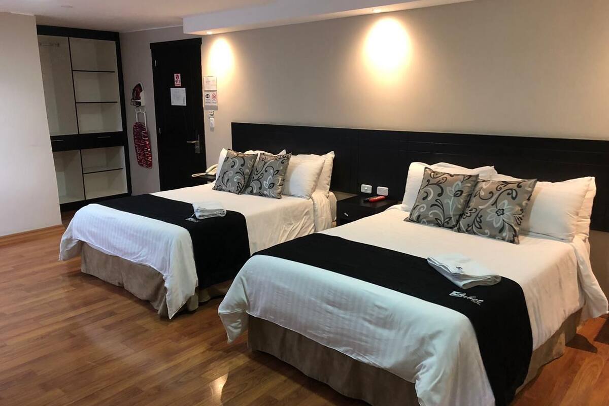 A hotel room with two beds, each with a black headboard, white bedding, and decorative pillows. There is a wooden floor and a small table with a lamp.