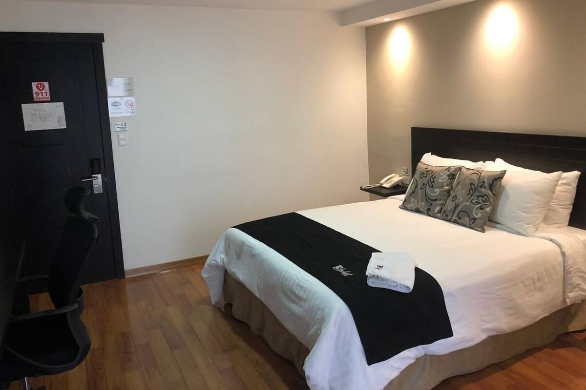 A hotel room with a bed, a desk, and a chair. There is a door with a sign on it.