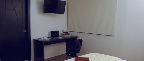 A hotel room with a bed, a desk with a laptop, a TV, and a window with a blind.