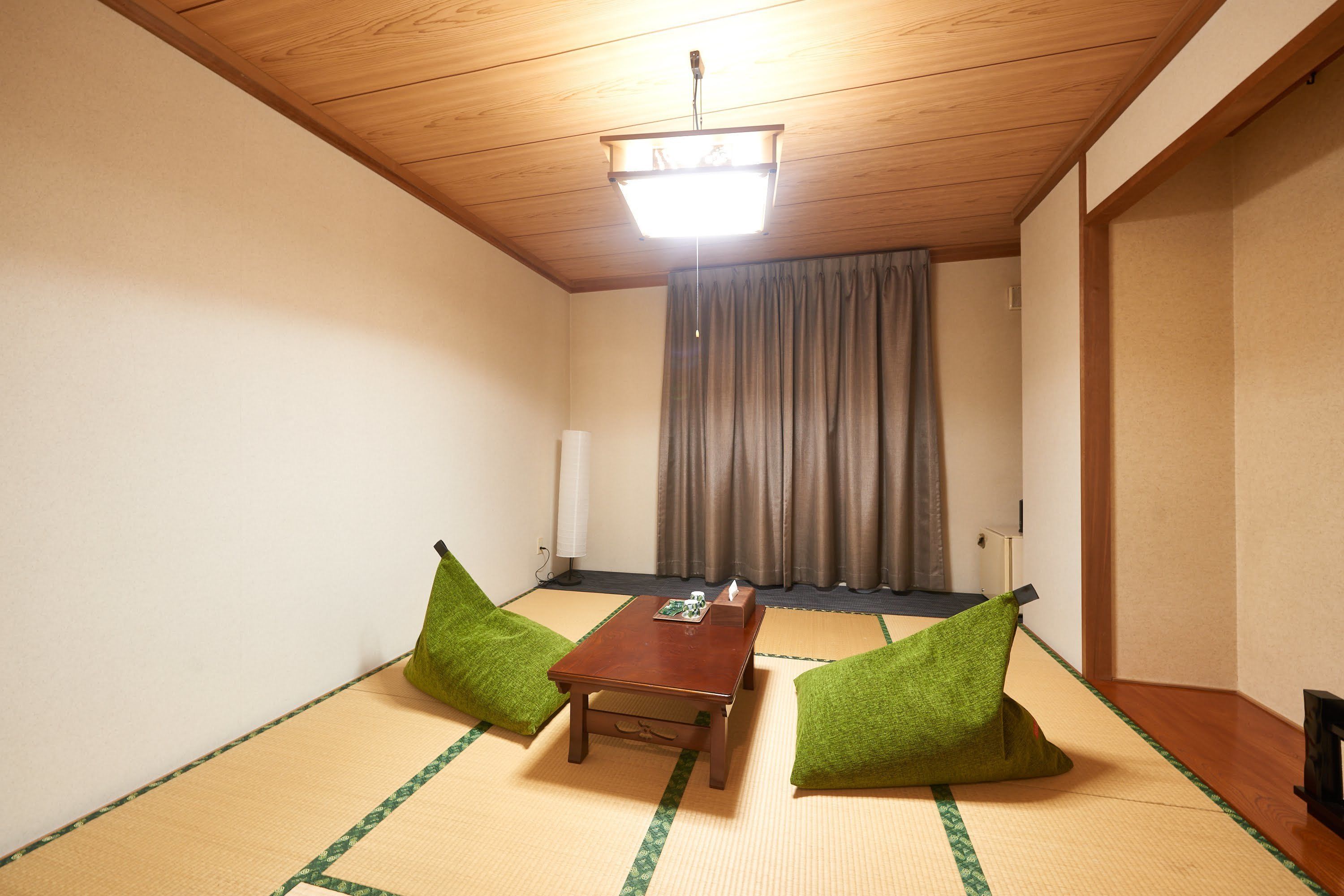 Una habitación con suelo de tatami, techo de madera, una mesa con cojín verde y una cortina.